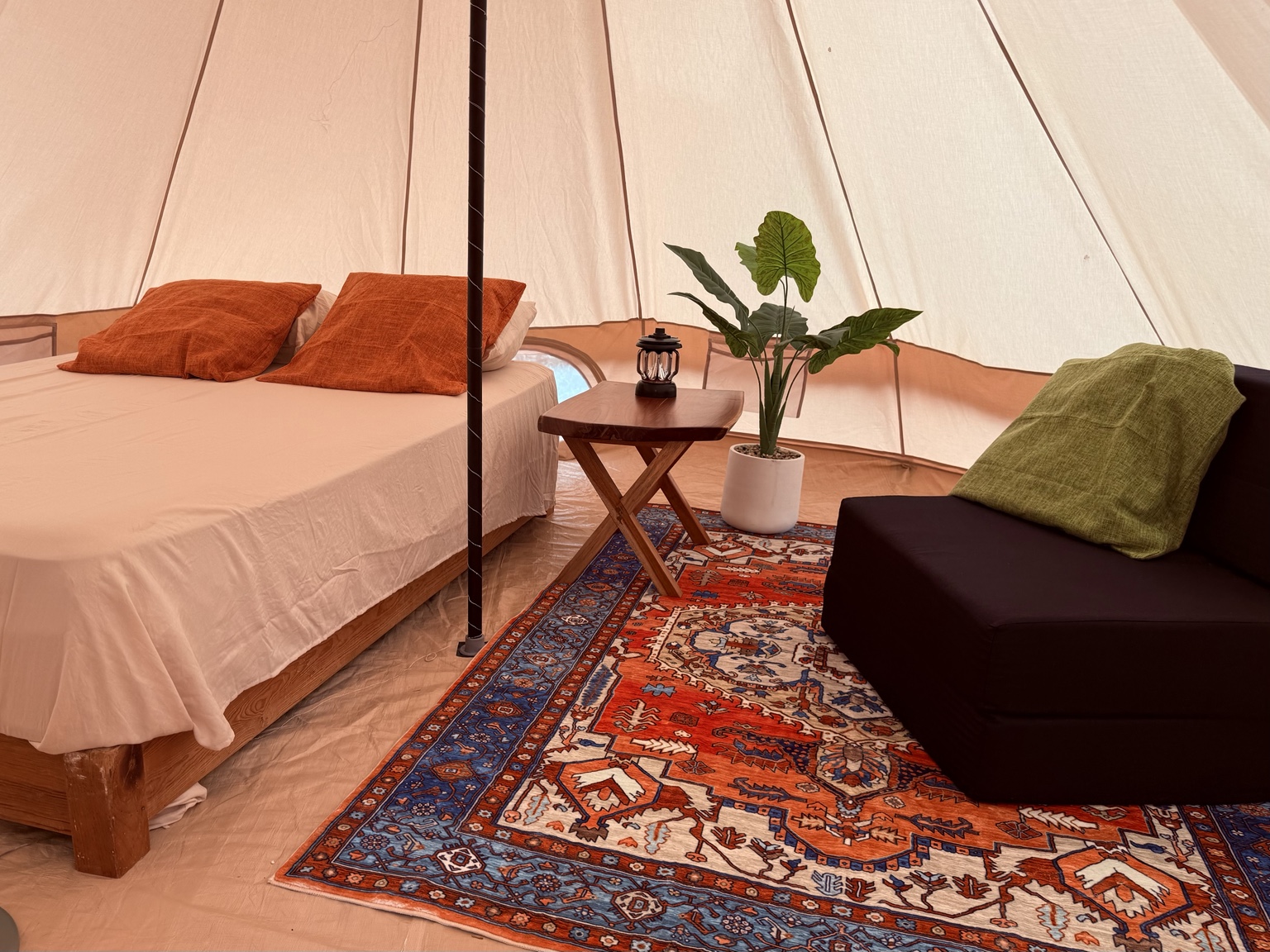 Glamping tent interior with bed, rug, plant, lantern, and futon.
